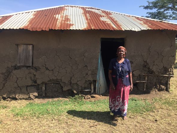 An old woman dressed in a blue top and maroon skirt. She is standing in front of a house. The houses has dilapidated walls and the door is open